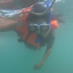 Snorkeller in a white-framed mask and orange life jacket looking at the camera underwater while holding the float ring rope