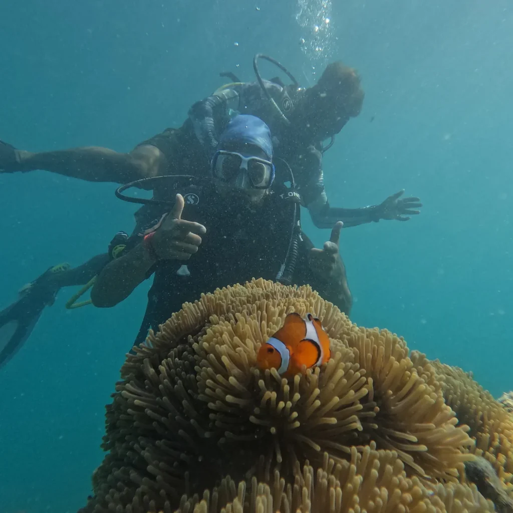 Diver giving a thumbs up next to a clownfish in its anemone while scuba diving at Nemo Beach, Havelock