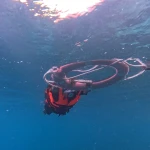 Snorkeller in an orange life jacket hanging under the float ring in deep blue water, seen from below