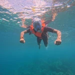Snorkeller in a full-face mask and orange life jacket swimming towards the camera with arms spread, reef below