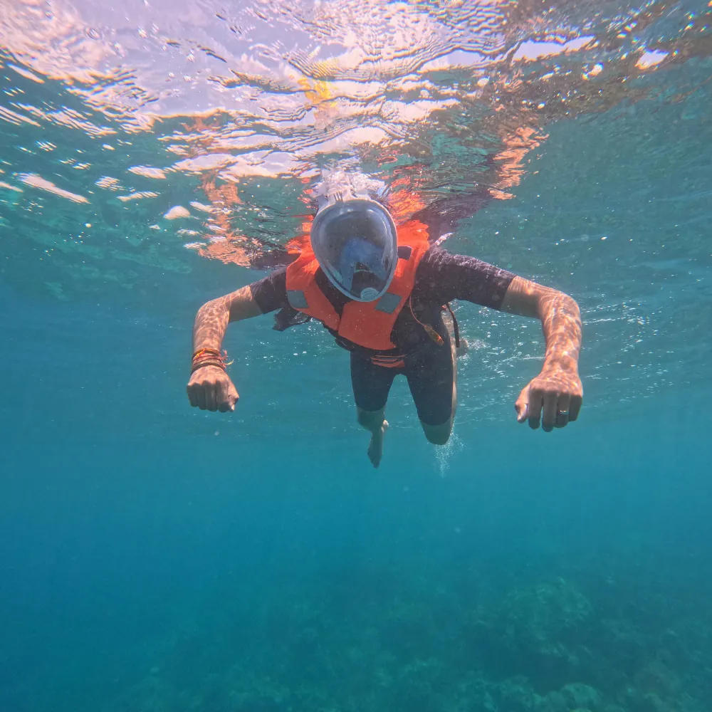 Snorkeller in a full-face mask and orange life jacket swimming towards the camera with arms spread, reef below