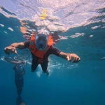 Snorkeller in a full-face mask and orange life jacket at the surface with a guide swimming below her