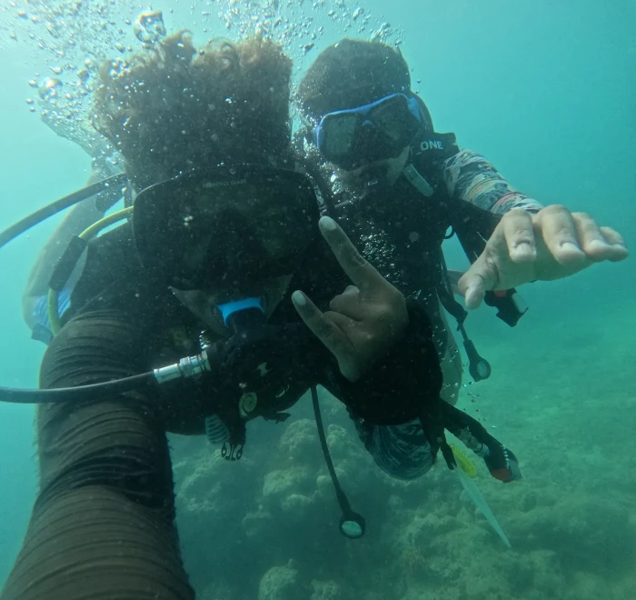 Two divers close to the camera, one giving a shaka sign underwater